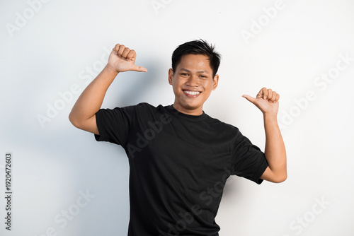 Photos Smiling young Asian man in a black shirt points both thumbs at himself against a white background, conveying confidence, pride, self-esteem, and playful affirmation