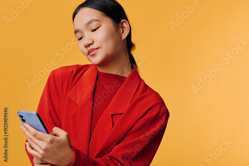 Fotografi Young woman in red jacket holding and looking at smartphone with slight smile on yellow background