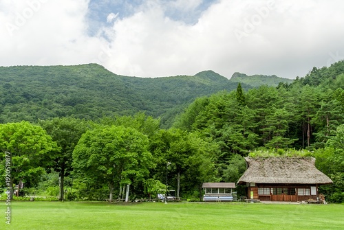 西湖 野鳥の森公園の夏：緑の芝生と茅葺き民家を包む雄大な山並み / Summer at Saiko Wildbird Forest Park: Majestic Mountains Embracing the Green Lawn and Thatched House