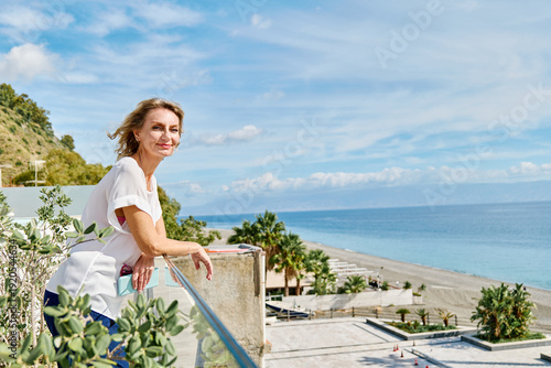 Woman traveler looking at camera enjoying the sun, admire seacoast view and breathing fresh sea air in hotel terrace.
