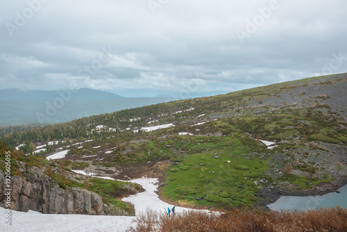 Two tourists on snowfield on hillside and hill silhouettes far away in low clouds. Scenic top view from glacier on steep slope to hilly terrain with turquoise lake under cloudy sky in rainy weather.