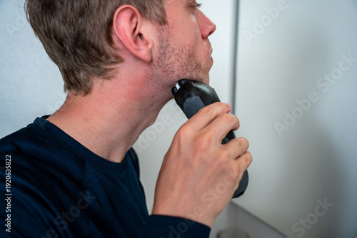 Young man using modern electric shaver in bright white bathroom for morning grooming routine. Close up of male shaving with electric razor in clean bathroom interior.