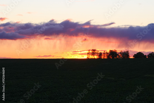 Dramatic sunset clouds over rural horizon with silhouetted trees and glowing sky. Sunlight filters through rain curtains creating calm dramatic mood in open countryside, concept of weather change