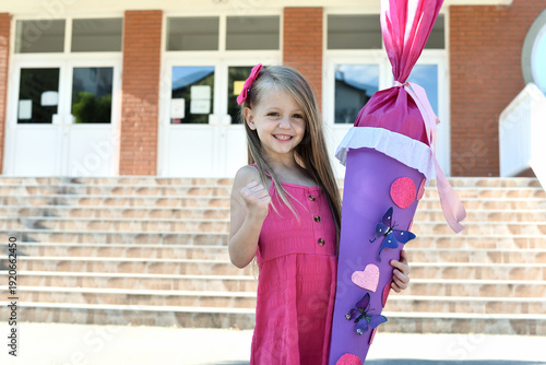 Happy little schoolgirl in a pink dress standing on the stairs of an elementary school, smiling and holding a large purple handmade school cone (Schultüte) decorated with hearts and butterflies on her
