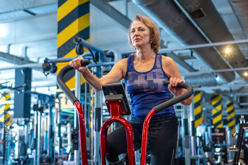 Foto Senior woman riding air bike in modern gym, smiling during cardio workout, active lifestyle and healthy aging