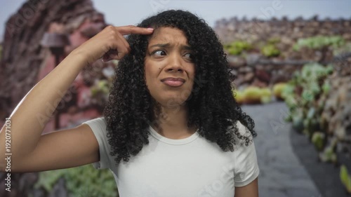 African american woman wearing white tshirt points finger to head in green park; uncertainty hesitation introspection.