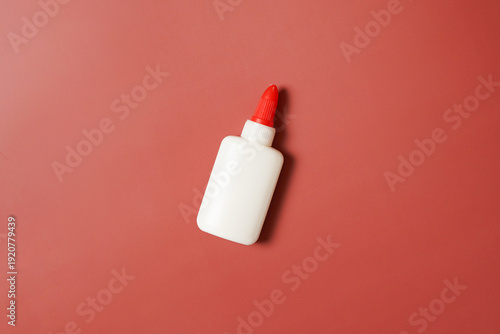 Top-down view of a single white glue bottle with a red applicator cap. Minimalist flat lay composition on a solid reddish background. Ideal for school, crafts, DIY, and office supplies.