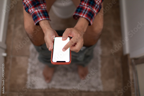 Картината върху платно Young man sitting on the toilet while texting and using phone scrolling social m