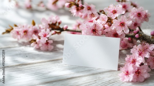 A branch of cherry blossoms rests on a light surface with a blank card positioned nearby. The flowers are pink and vibrant signifying the arrival of spring.