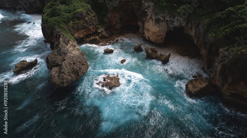 The scene shows waves crashing against large rocks along a shoreline in Bali. The ocean water is blue and clear. Lush greenery surrounds the rocky area.
