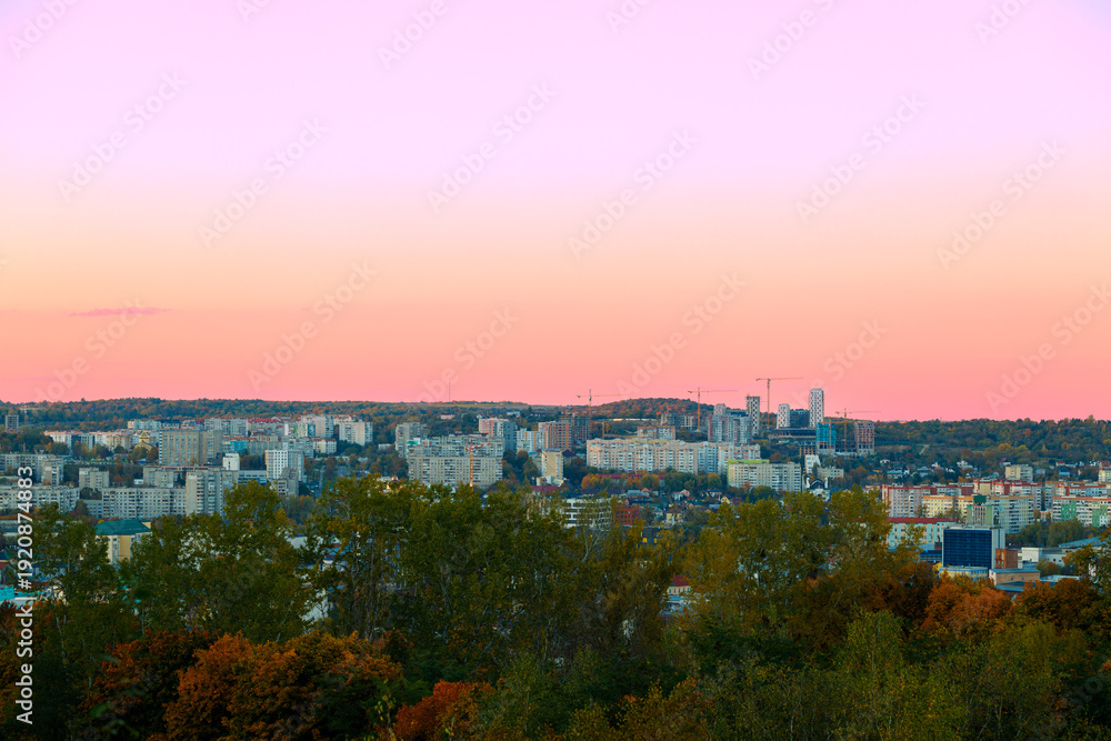 Fototapeta premium Panoramic cityscape at sunset with pastel sky and lush green trees in the foreground