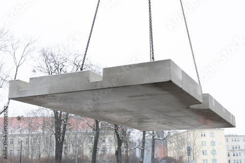Large prefabricated concrete slab lifted by crane during installation process. Heavy precast element suspended in air at an urban construction site.