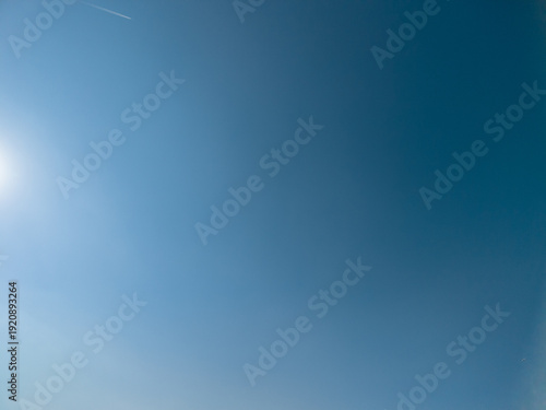 Expansive view of a bright clear blue sky, featuring a glowing sun flare in the corner and a faint airplane contrail tracing across the upper backdrop, offering copy space for various designs