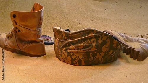 An arrangement of abandoned military gear, including a worn out boot, helmet, and shattered protective eyewear, partially buried in desert sand, symbolizing the harsh realities of conflict.