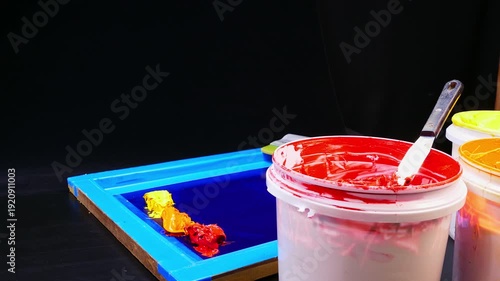 A bright studio setup featuring open buckets of vibrant inks beside a blue screen printing frame. The mix of tools and hues reflects the artistry and preparation behind professional printmaking.