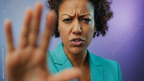 A serious African American woman in a teal blazer makes a stop gesture with her hand, expressing rejection or denial against a purple and blue studio background.