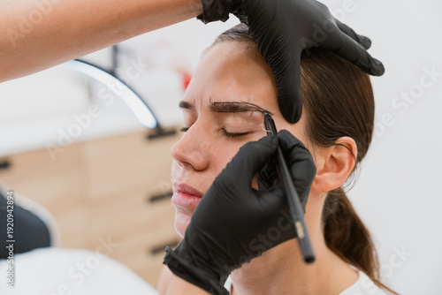 Beautician drawing eyebrow shape on young woman face with pencil