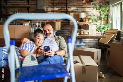 Photos Smiling mature couple on couch using smartphone at new home during move
