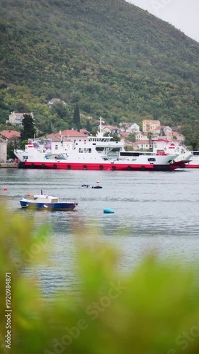 White and red car ferry loading passengers at dock with mountains and a small town. Bay of Kotor, Montenegro