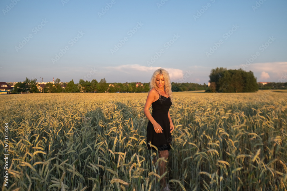 Fototapeta premium Female model with curly blonde hair wearing a black dress stands in a field of wheat during sunset, with soft clouds and a colorful sky in the background