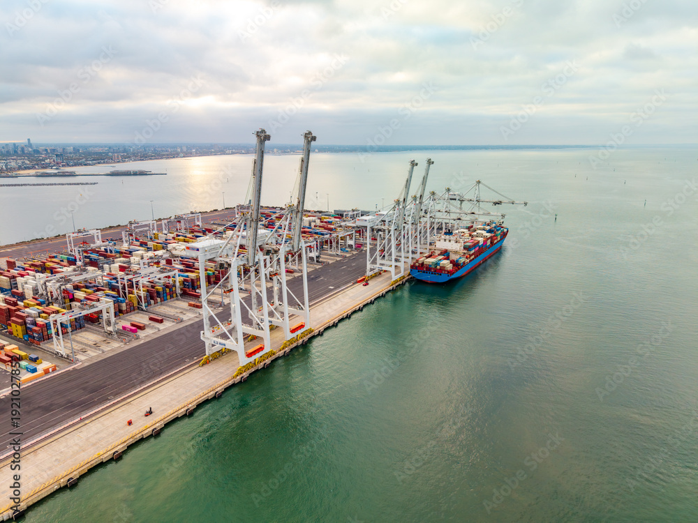 Fototapeta premium Aerial view of a busy container ship docked at the Port of Melbourne, with gantry cranes actively handling stacks of colorful shipping containers.