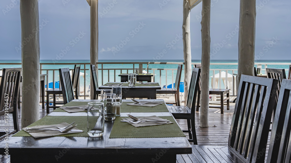 Fototapeta premium Seaside cafe. The tables are set. There are cutlery and glasses on the napkins. Wooden chairs. Behind the latticed railings is an aquamarine ocean. Sun umbrellas on the beach. Cuba. Varadero. Resort. 
