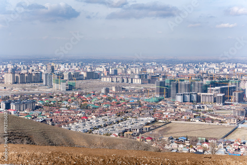 A high-angle, wide shot of a developing city skyline in Bishkek, Kyrgyzstan, where modern high-rise buildings under construction contrast sharply with older, low-rise residential areas in the foregrou