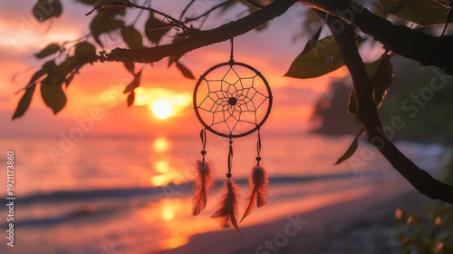 A dreamcatcher hangs from a tree branch over a beach at sunset. The dreamcatcher is decorated with feathers and is suspended from a branch. The scene is serene and peaceful