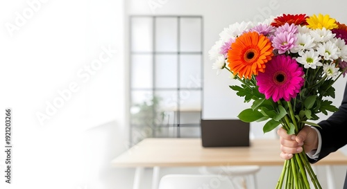 Person holding a bouquet of colorful flowers in a modern office setting with a laptop on a wooden desk