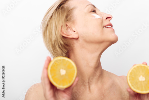 Mature woman with short blonde hair holds two halves of fresh orange in hands, applying white cream on face. Concept of natural skincare, vitamin c, healthy lifestyle and beauty care.