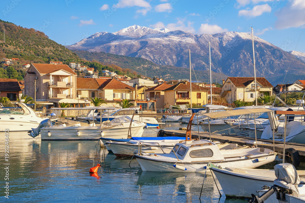 Obraz premium Fishing boats in harbour. Mediterranean landscape on sunny winter day. Montenegro. View of marina Kalimanj in Tivat city. Snow-capped Lovcen mountain