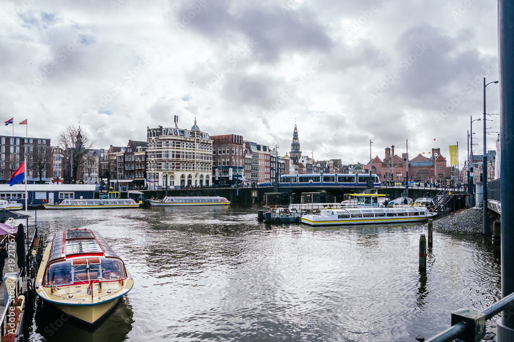 Fototapeta premium Amsterdam canal scene with tour boats cruising past historic waterfront buildings and a tram crossing a bridge under a cloudy sky, capturing urban travel and sightseeing