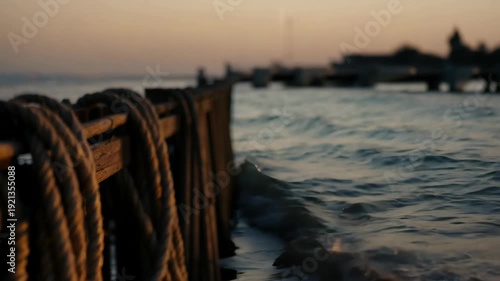 Coiled ropes on a wooden pier edge with waves lapping against it during sunset, creating a tranquil coastal scene with blurred background elements