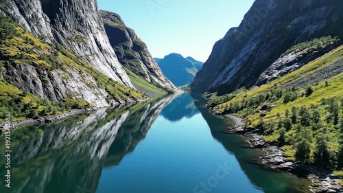 Narrow fjord with steep cliffs and vibrant greenery reflects in tranquil blue waters under a clear sky, showcasing Norway's stunning natural landscape