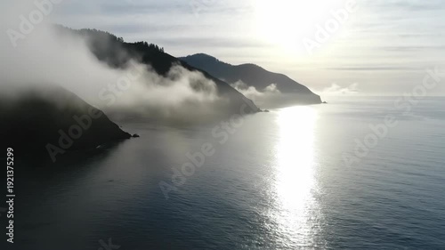 Coastal scene features fog enveloping mountains while sunlight reflects on the ocean, creating a peaceful atmosphere with natural elements in harmony