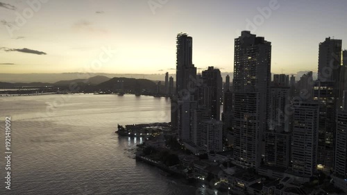 Cinematic aerial drone footage of the sunset over Cinta Costera in Panama City. The golden light of the sunset illuminates the coastal skyline, oceanfront boulevard, and modern buildings of the city, 