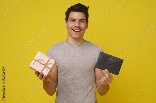 A young man with a mustache smiles happily as he holds out a wrapped gift box and a blue envelope. He is standing in front of a solid yellow backdrop