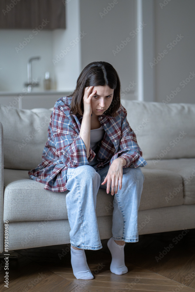 Fototapeta premium Anxious stressed young woman holding head with tense, lost in depressed thoughts. Worried female sits alone on sofa, touching temple with hand. Mental fatigue, loneliness, inner conflict, frustration.