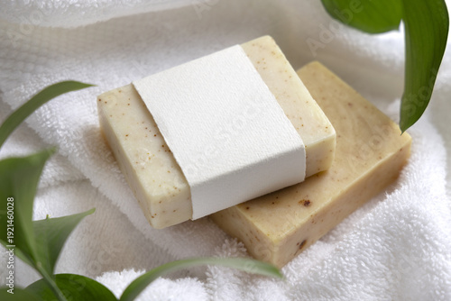 Mockup of beige solid soap bar with unbranded paper label near green leaves on white bath towels