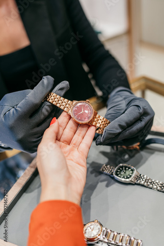 A professional jeweler wearing grey gloves presents an elegant rose gold watch with a pink dial to a customer during a luxury retail shopping experience.