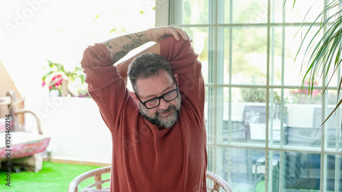 Obraz na plátně Active mature bearded man exercising inside home with stretching exercises for shoulders and arms in a modern living room