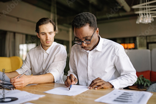 Two men, white and black, working together at a wooden table in a contemporary office, one is taking notes while the other reviews documents, both engaged in a productive discussion.