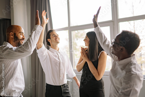 Four diverse office workers high-fiving and celebrating success indoors in light office