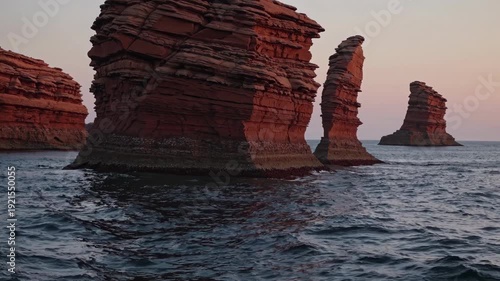 Rock formations emerge from calm waters at sunset, showcasing unique geological structures with layered textures and vibrant colors against a serene coastal backdrop