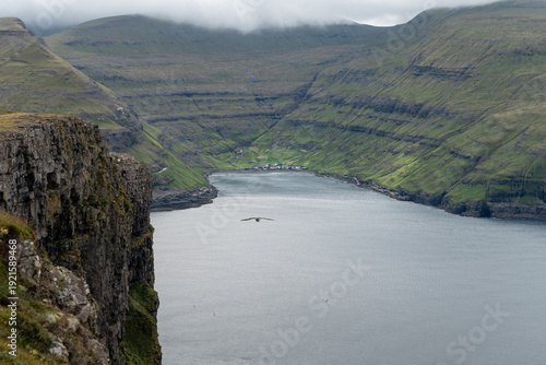 A panoramic view of the secluded village of Tjørnuvík nestled along a quiet bay and surrounded by dramatic mountains viewed from a high-altitude vantage point
