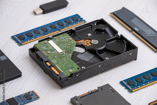 Opened hard disk drive with visible platter and circuit board surrounded by RAM modules and SSD on wooden desk