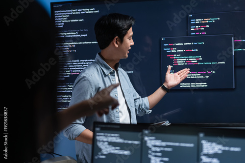 Photos Asian Male software engineer pointing at source code on a large screen during a tech team meeting, explaining Code on the Screen, Discussing on coding task, New software development project in office