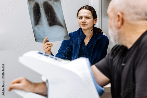 Man having his blood pressure measured with a digital sphygmomanometer in medical facility while doctor reviews lung X-ray in background. Medical examination concept

