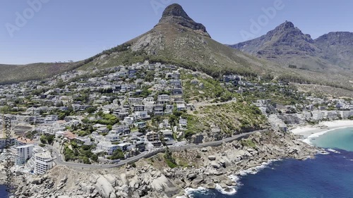 Drone flies left from Sea Point to Clifton Beach with view of Lion's Head and Table Mountain on sunny day in Cape Town, South Africa
