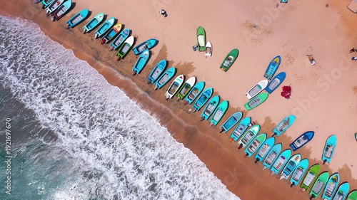 Aerial view of colorful fishing boats lined up along a sandy beach with waves from the Indian Ocean at Nilaveli, Sri Lanka. No people visible.
